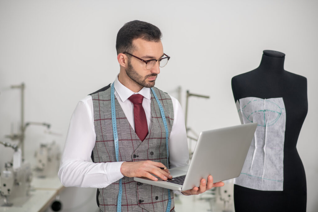 Fashion design studio. Male tailor standing, holding laptop, typing something, thinking