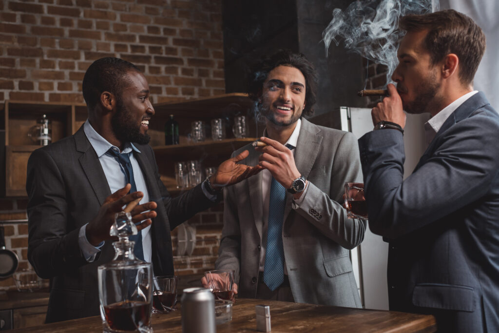 smiling multiethnic male friends in suits talking, drinking whiskey and smoking cigars in a ciagr lounge